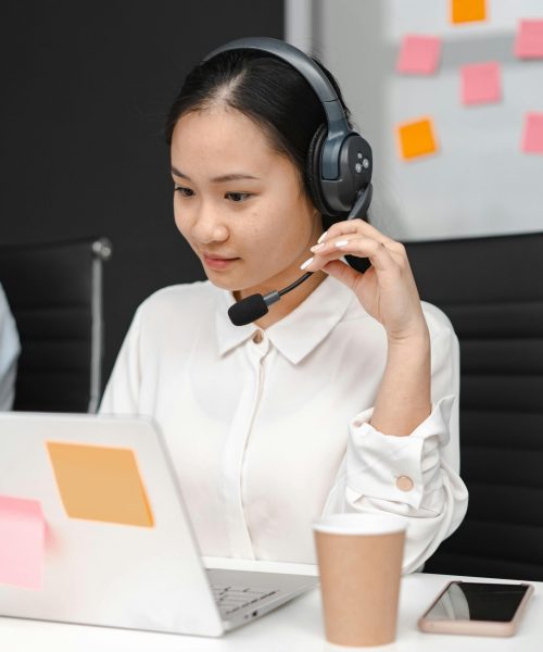Asian woman using a laptop and headset for customer support in an office environment.